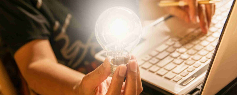 A person on a laptop holding a lit lightbulb